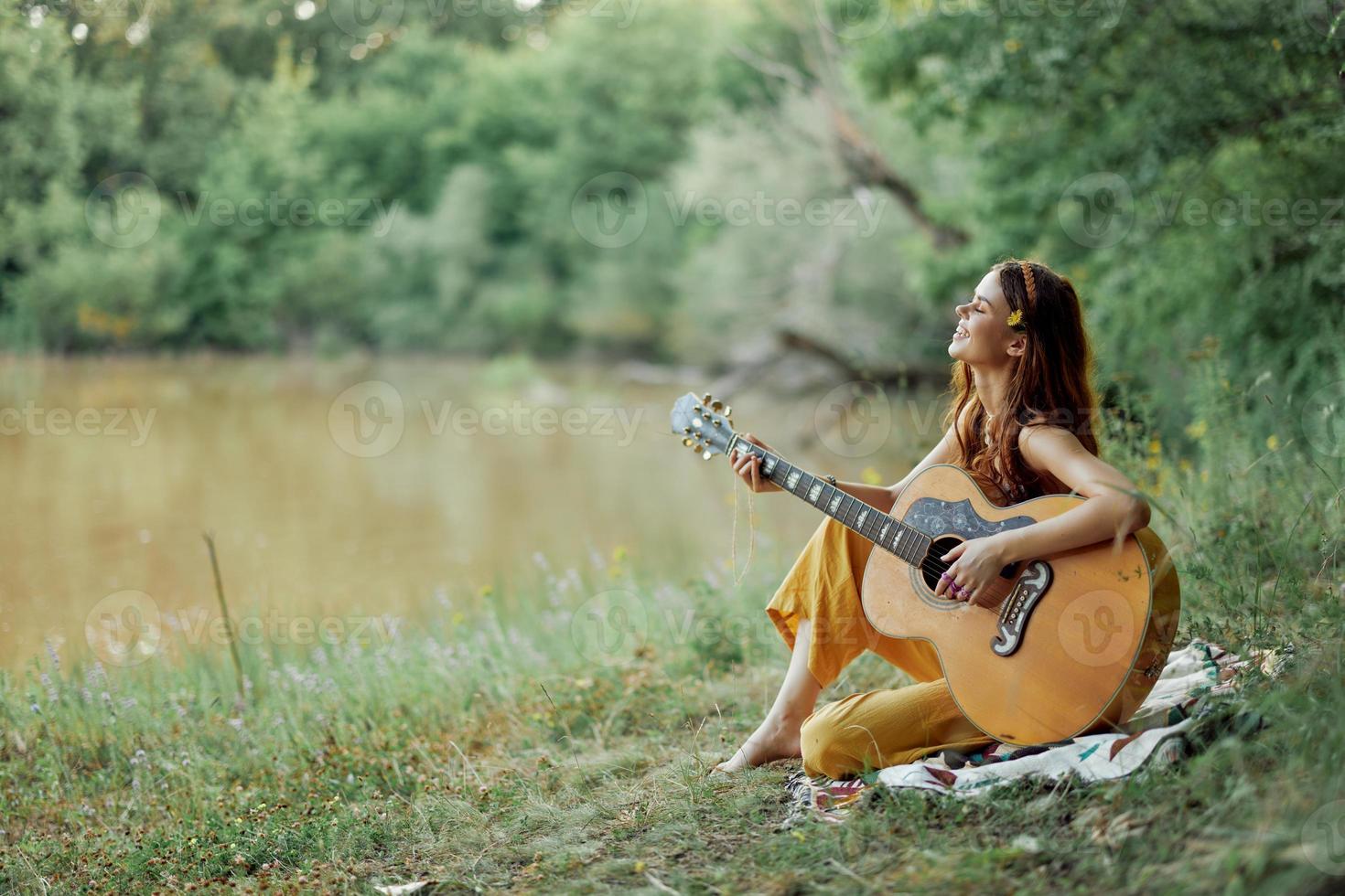 hippie kvinna spelar gitarr leende och sång låtar i natur Sammanträde på en pläd förbi de sjö i de kväll i de strålar av de miljö Sol. en livsstil i harmoni med de kropp och natur foto