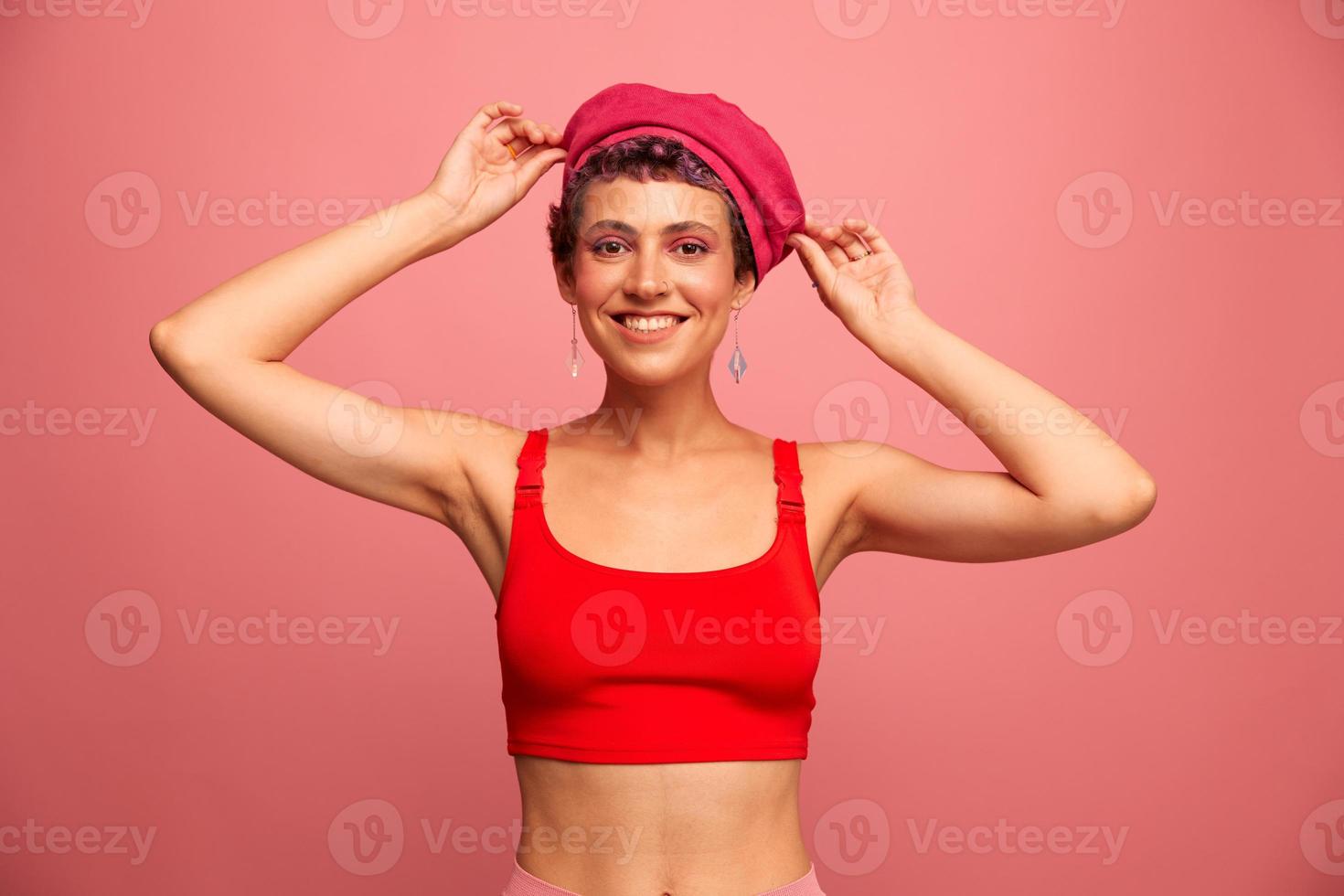 ung atletisk kvinna med en kort frisyr och lila hår i en röd topp och en rosa hatt med ett atletisk figur ler och grimaser som visar tecken med henne händer ser på de kamera på en rosa bakgrund foto