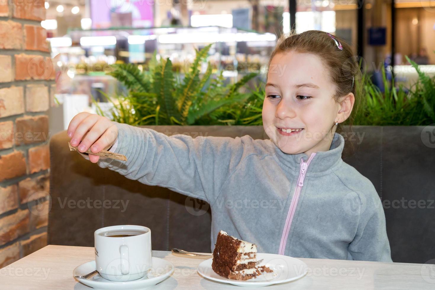 leende liten flicka i en Kafé på de tabell häller socker in i en kopp Nästa till en tallrik med kaka foto