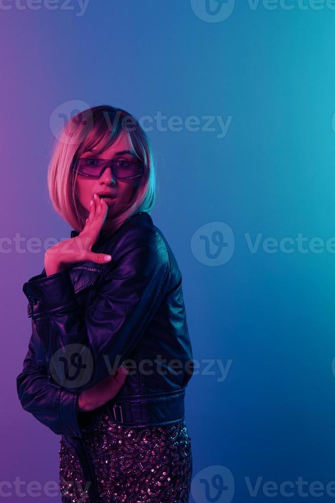 upphetsad chockade sexig skön blond kvinna i läder jacka glittrande klänning trendig solglasögon stänga mun med hand Framställ isolerat i blå rosa Färg ljus studio bakgrund. neon fest begrepp foto