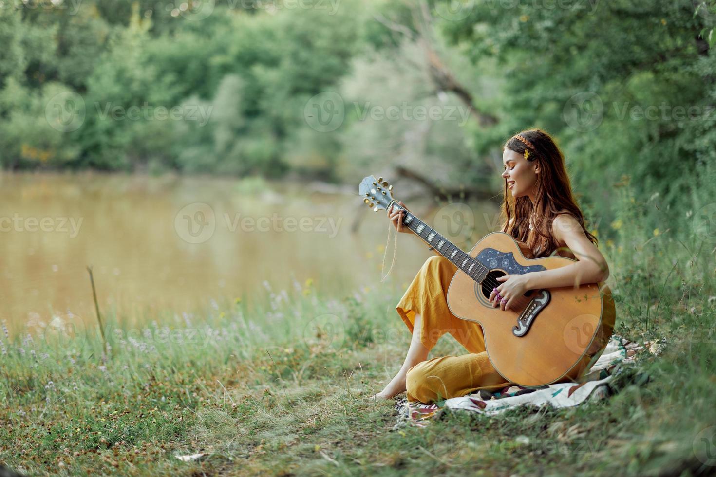 hippie kvinna spelar gitarr leende och sång låtar i natur Sammanträde på en pläd förbi de sjö i de kväll i de strålar av de miljö Sol. en livsstil i harmoni med de kropp och natur foto