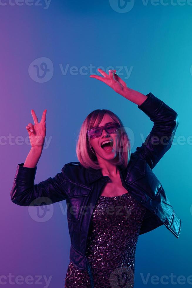 haft sexig skön blond kvinna i läder jacka glittrande klänning trendig solglasögon dansa håll händer upp visa v-tecken Framställ isolerat i blå rosa Färg ljus studio bakgrund. neon fest begrepp foto