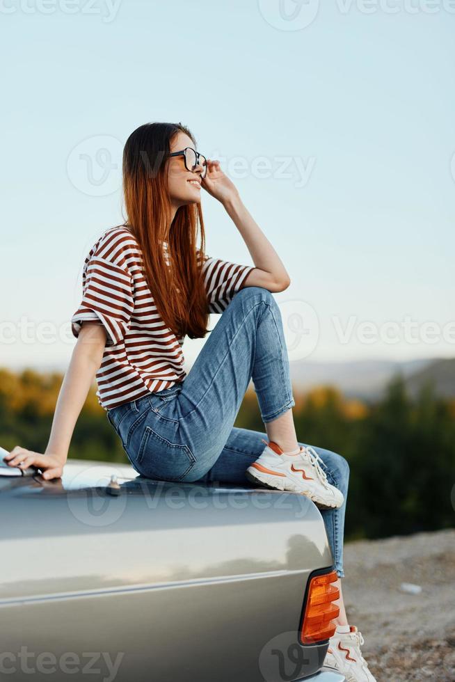 en mode kvinna i eleganta solglasögon, en randig t-shirt och jeans sitter på de trunk av en bil och utseende på de skön natur av höst. resa livsstil foto