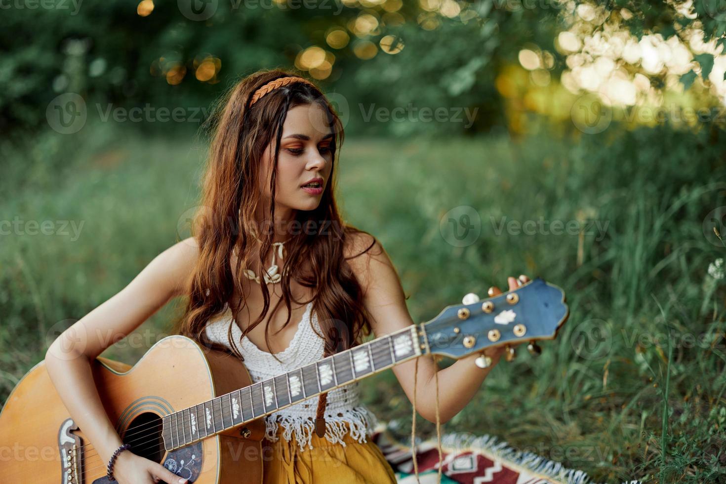 flicka hippie kvinna spelar gitarr i miljövänlig kläder Sammanträde på de jord utanför i natur i de falla tittar på de solnedgång foto