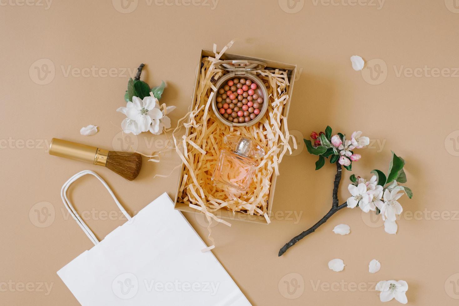 kosmetisk låda för en kvinna rodna, borsta och parfym på en beige bakgrund omgiven förbi delikat vit blommor. platt lägga foto