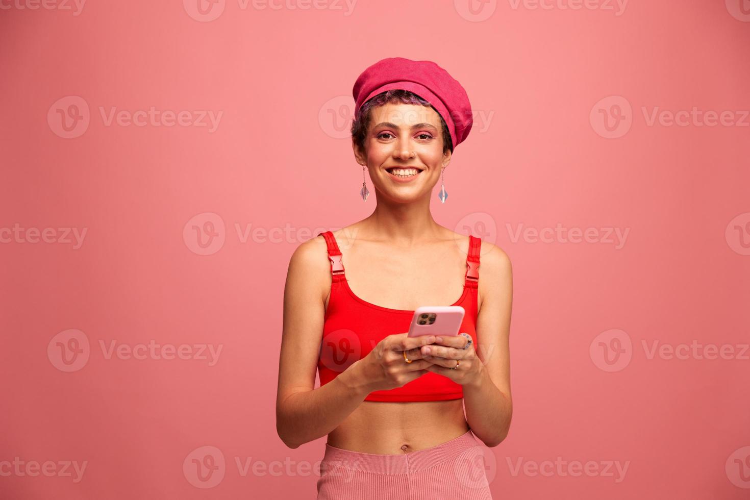 ung kvinna bloggare med färgad rosa hår och kort frisyr vända genom de telefon skärm och skriver en meddelande med en leende och överraskning i eleganta kläder på en rosa bakgrund foto