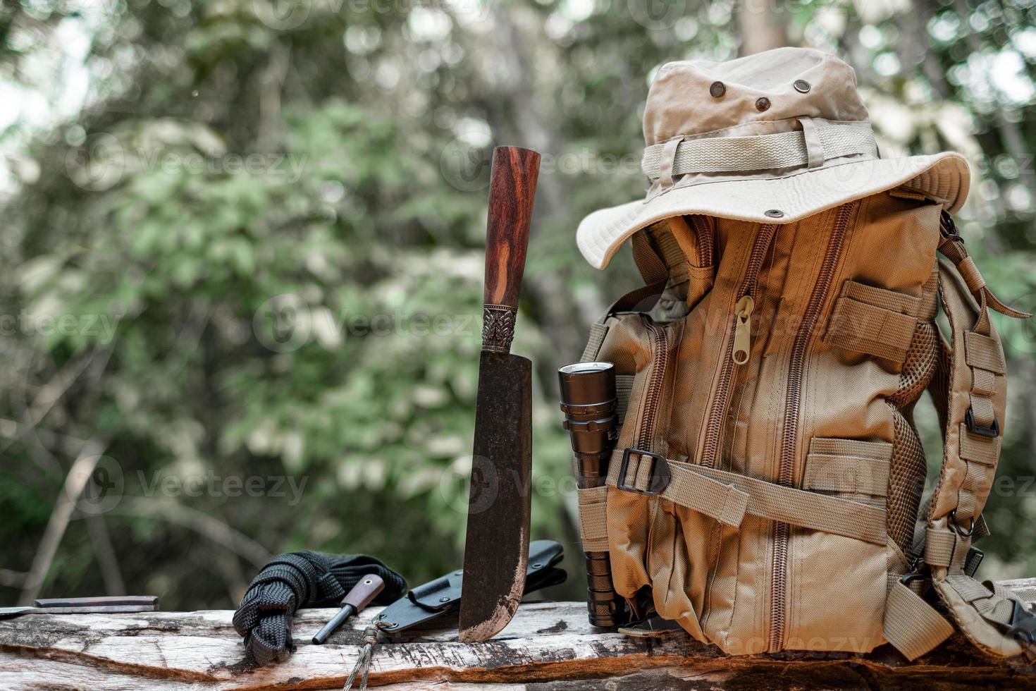 Utrustning för överlevnad hink hatt ryggsäck vandring kniv camping ficklampa vilar på trä- timmer i de bakgrund är en skog foto