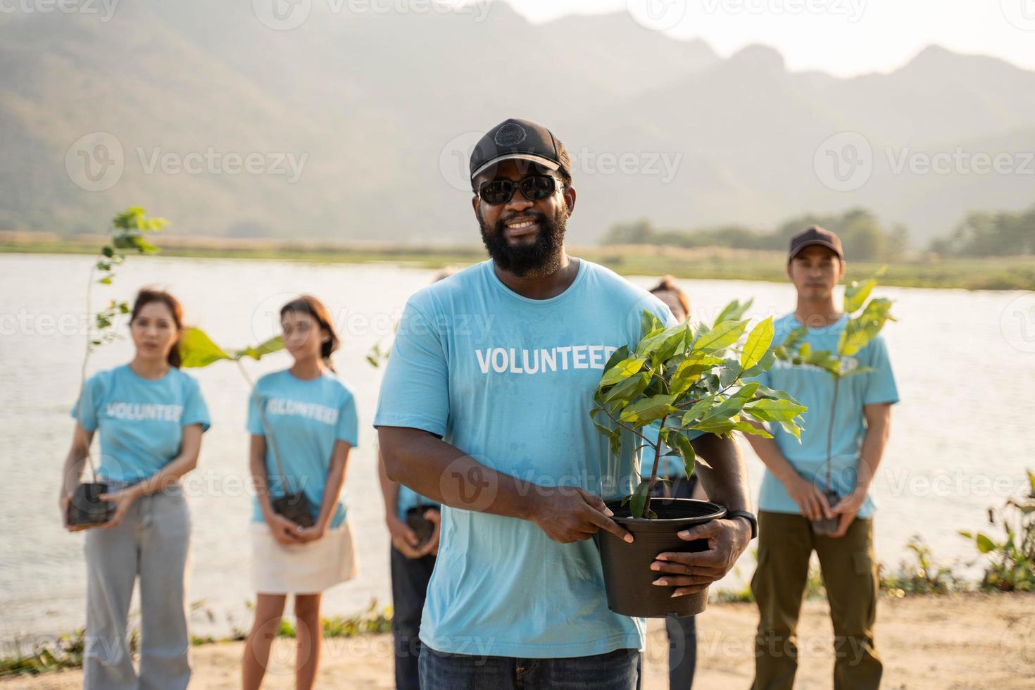 porträtt av afrikan man med bakgrund grupp av volontär- innehav pott med grön växt leende till kamera stående på flod. skydd av miljö och natur, ekologi begrepp. foto