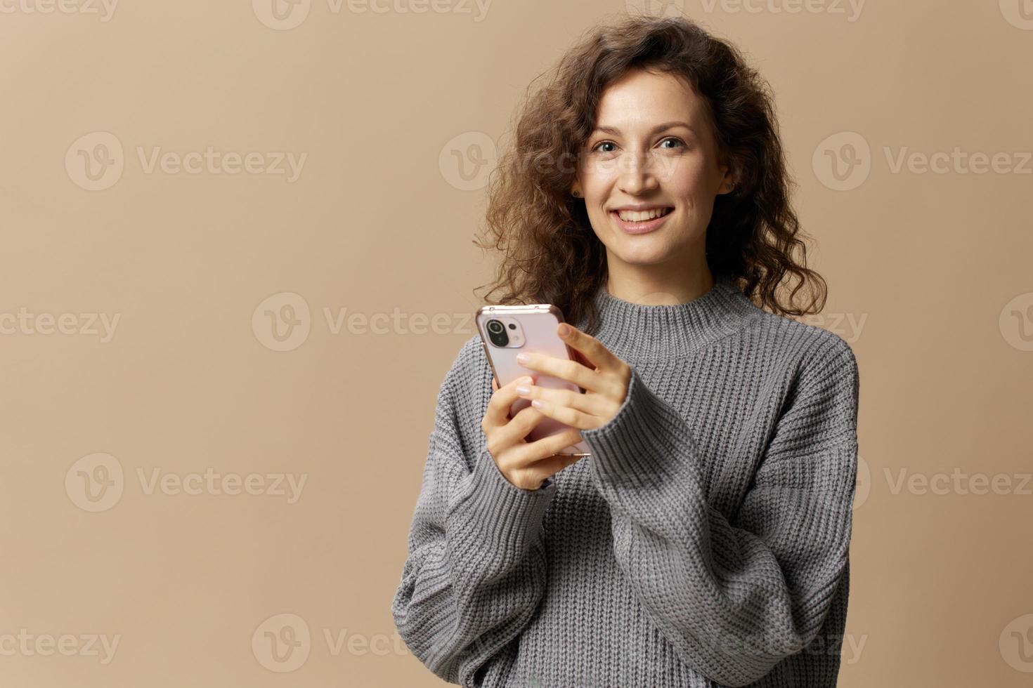 Lycklig lockigt skön lady i grå tillfällig Tröja chattar med pojkvän utseende på kamera Framställ isolerat på beige pastell bakgrund. social media, nätverk, distans kommunikation begrepp. kopia Plats foto