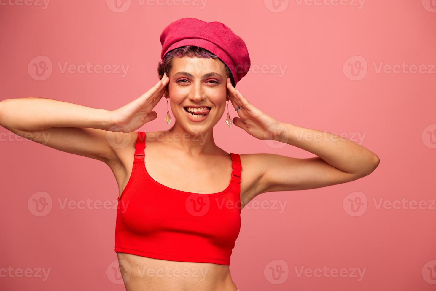 ung atletisk kvinna med en kort frisyr och lila hår i en röd topp och en rosa hatt med ett atletisk figur ler och grimaser ser på de kamera på en rosa bakgrund foto