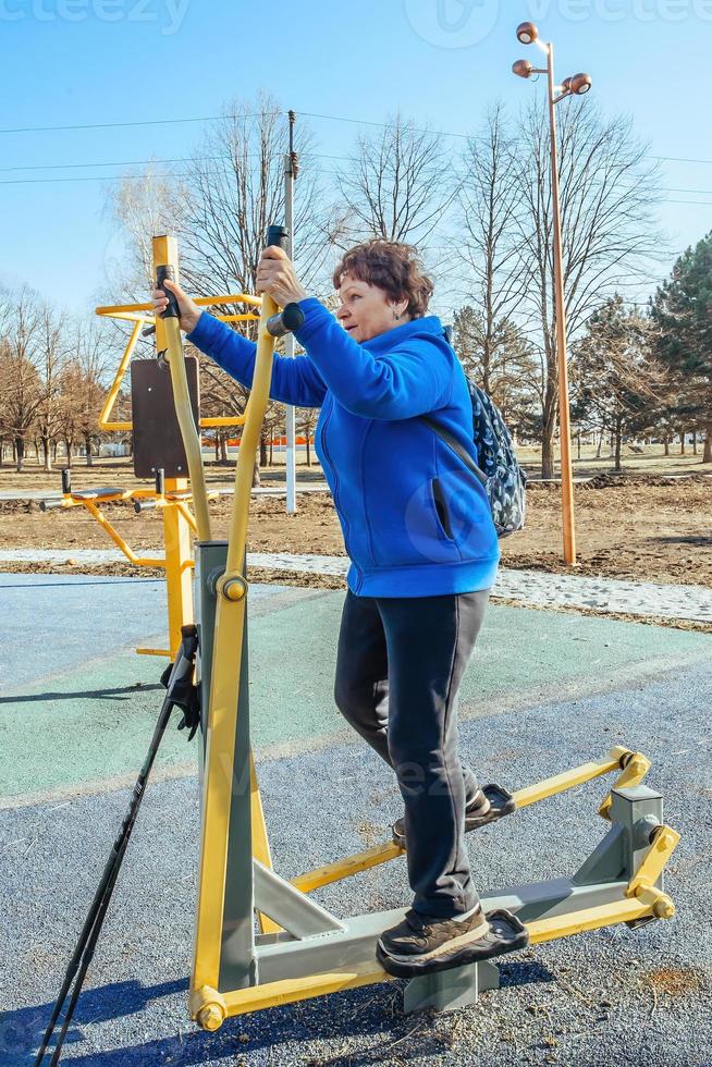 en pensionerad kvinna med en ryggsäck på henne axlar på ett aktiva promenad utomhus är engagerad i övning Utrustning i de parkera. senior kvinna tar vård av henne hälsa foto
