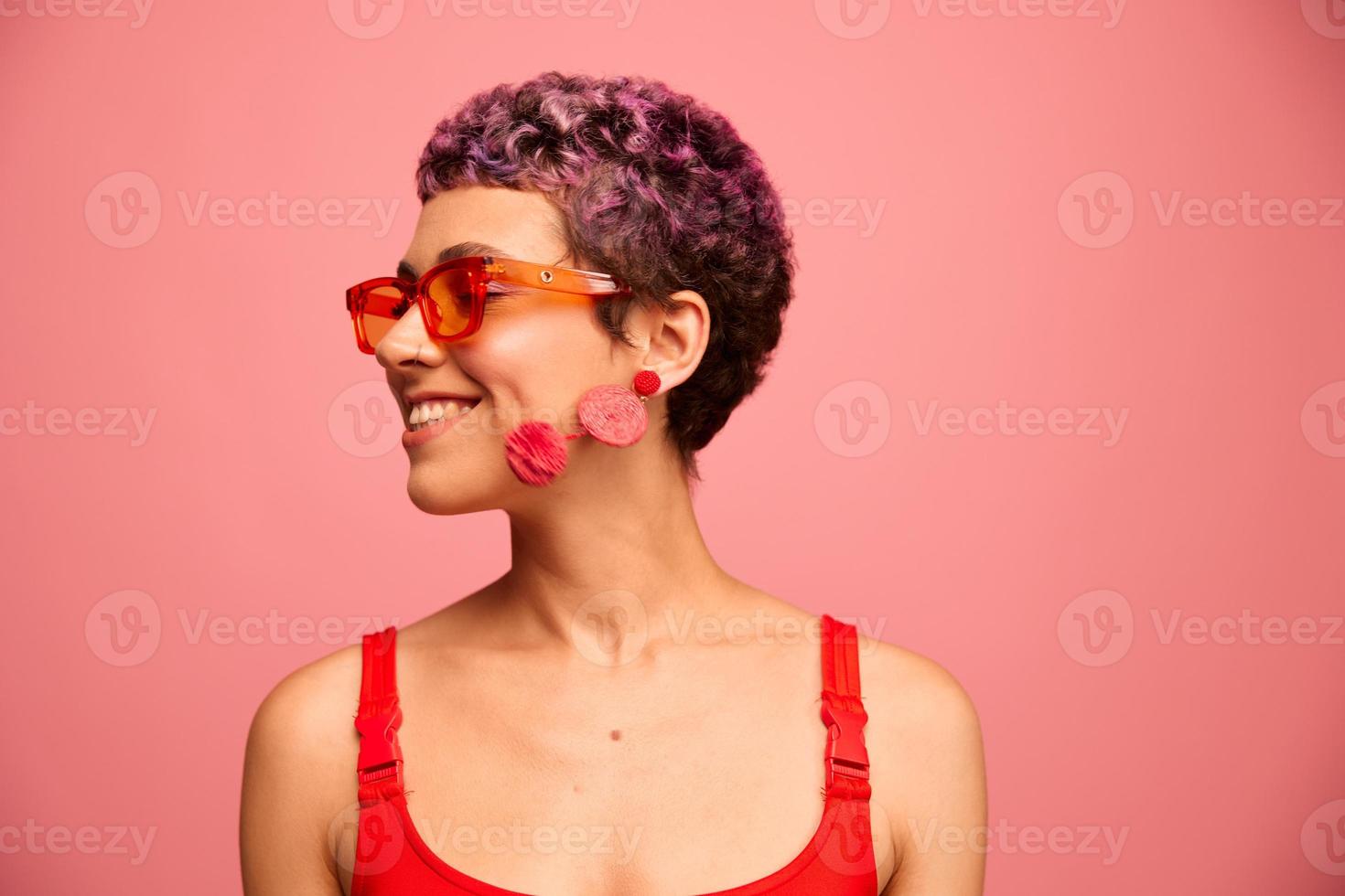 mode porträtt av en kvinna med en kort frisyr i färgad solglasögon med ovanlig Tillbehör med örhängen leende på en rosa ljus bakgrund foto