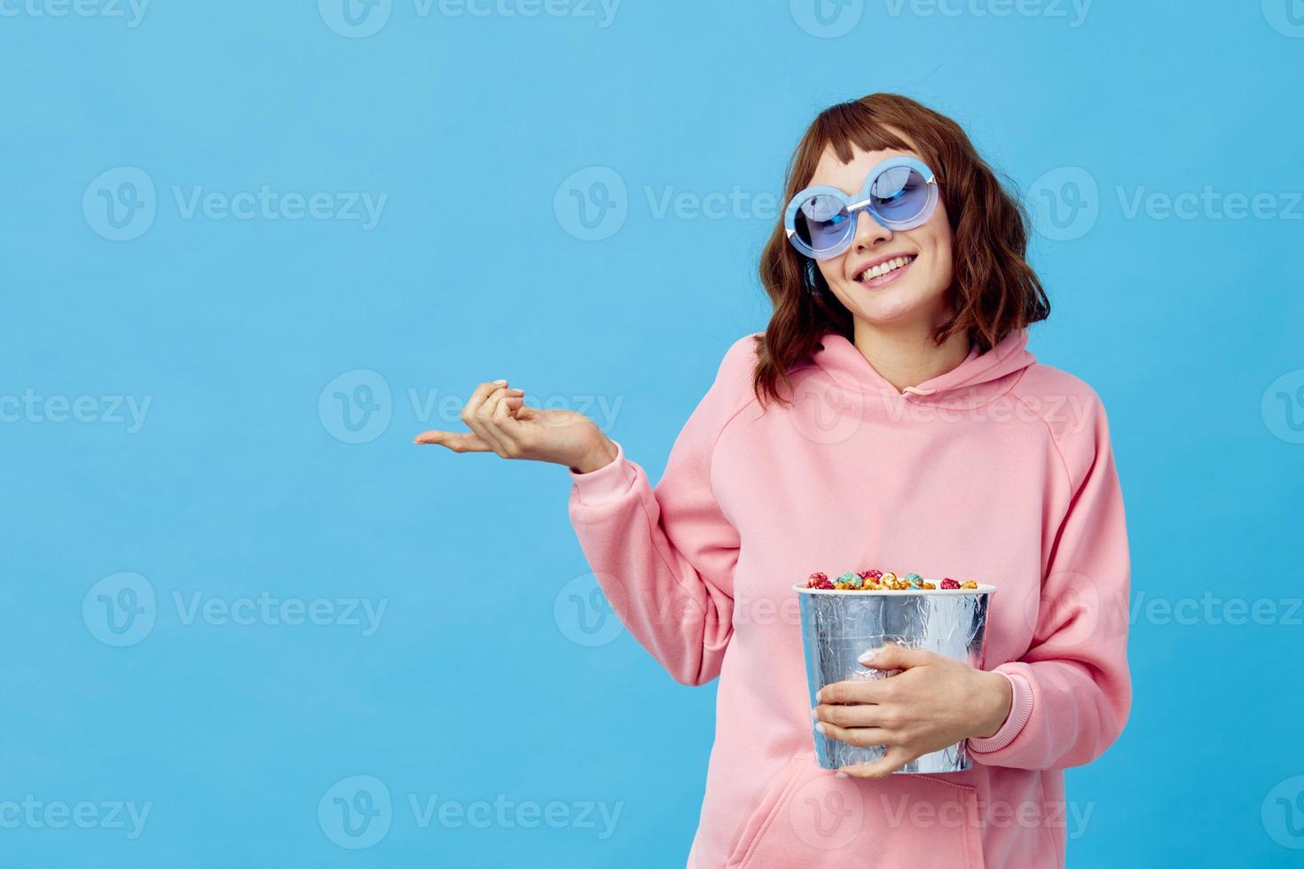 kväll film begrepp. härlig leende söt rödhårig lady i rosa luvtröja solglasögon med popcorn Framställ isolerat på blå studio bakgrund. kopia Plats baner. mode bio. underhållning erbjudande foto