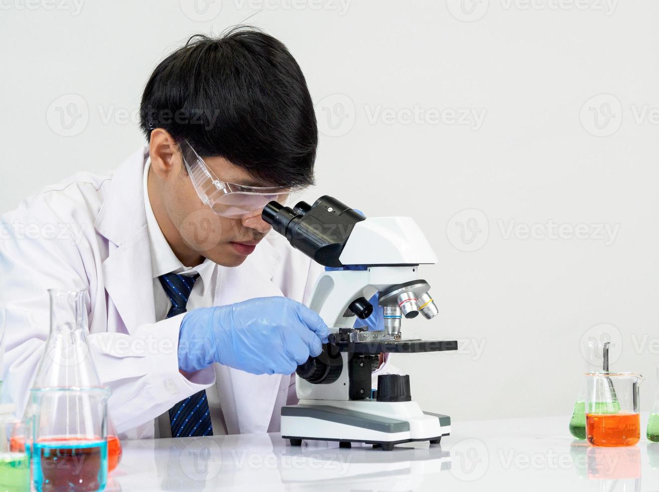 asiatisk manlig studerande forskare bär en läkares klänning i de labb ser hand på kemist. orsakade förbi blandning reagens i vetenskaplig forskning laboratorier med testa rör och mikroskop på de tabell foto