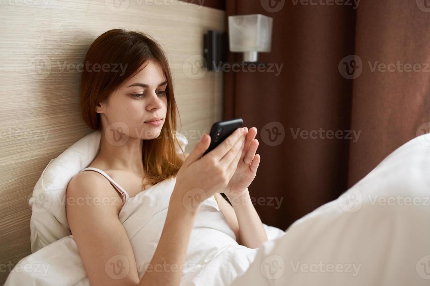 kvinna med mobil telefon lögner i säng nära fönster och gardiner design foto
