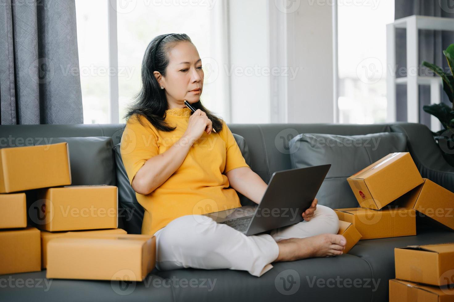 senior företag ägare kvinna förbereda paket låda och stående kolla upp uppkopplad order för leverera till kund på läsplatta, bärbar dator handla uppkopplad begrepp. foto