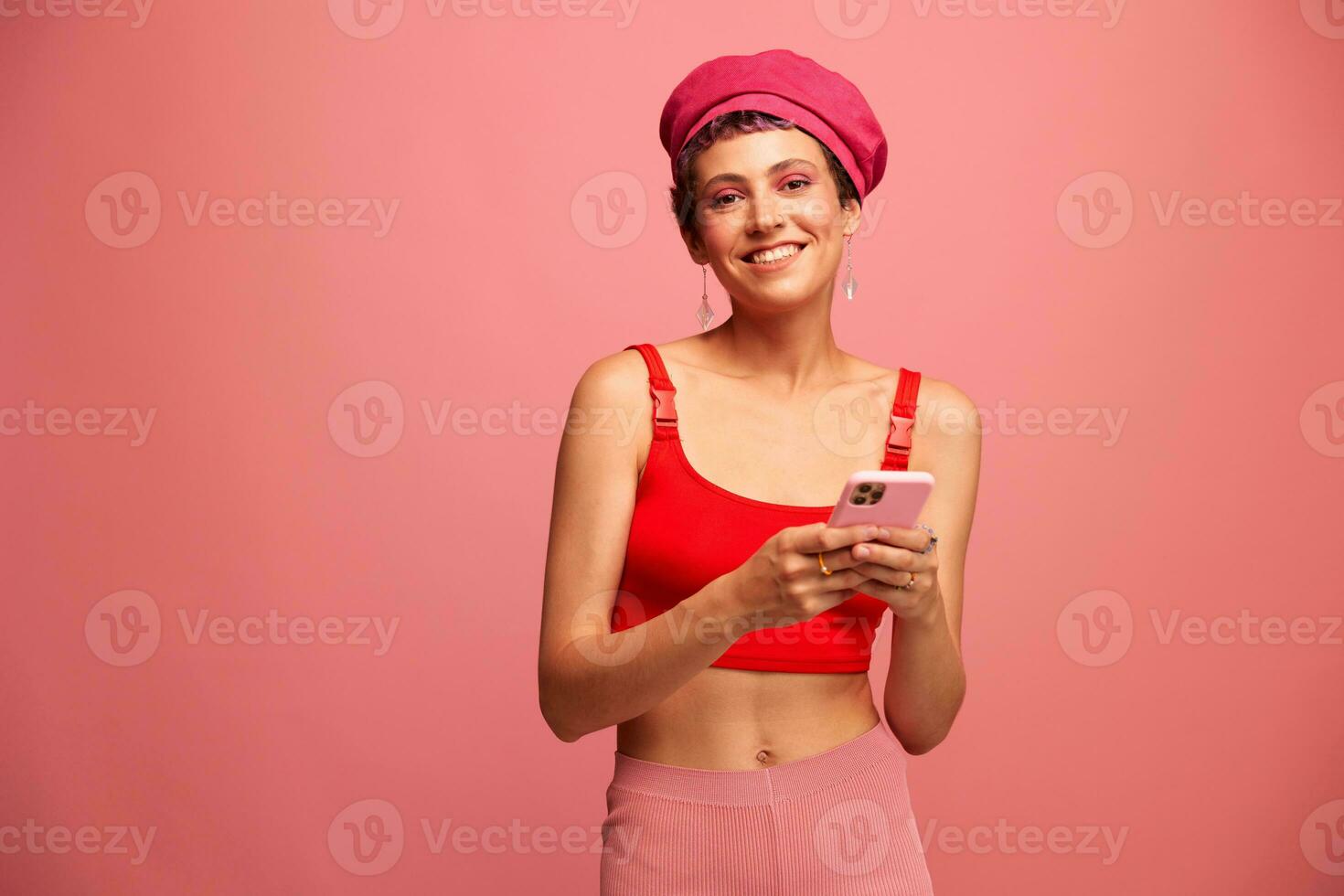 ung kvinna bloggare med färgad rosa hår och kort frisyr vända genom de telefon skärm och skriver en meddelande med en leende och överraskning i eleganta kläder på en rosa bakgrund foto