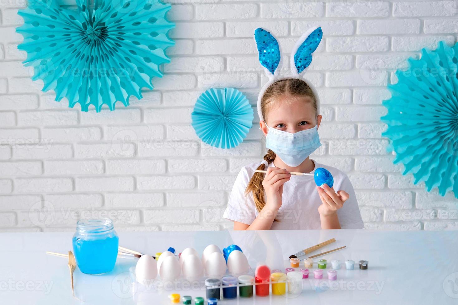 en liten flicka i en medicinsk mask målarfärger påsk ägg med en mönster. barn framställning påsk dekoration på Hem och bär ansikte mask till skydda från coronavirus foto
