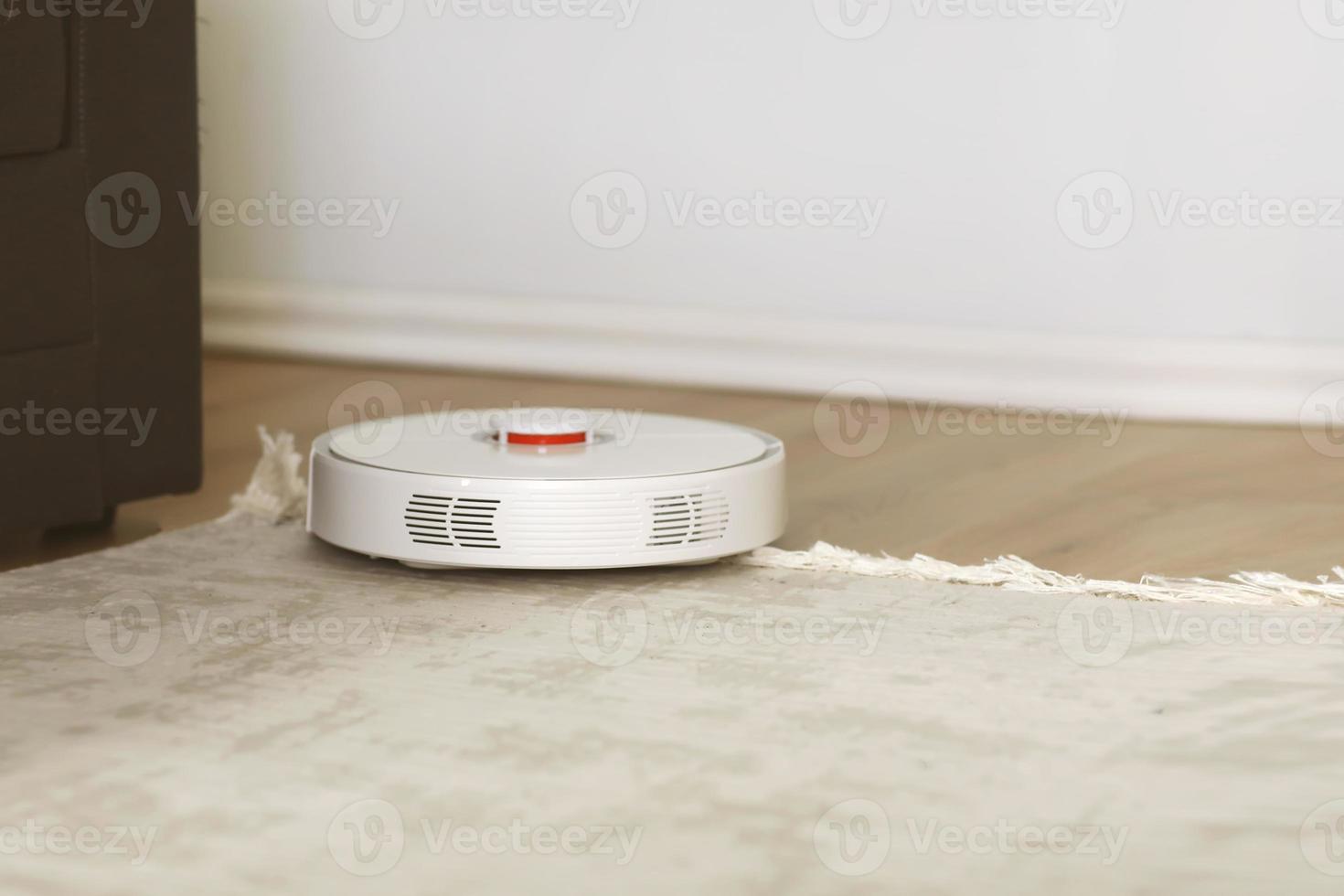 vit robot dammsugare på en matta rengöring damm i vardagsrum interiör. smart elektronisk hushållsteknik. grunt djup. foto
