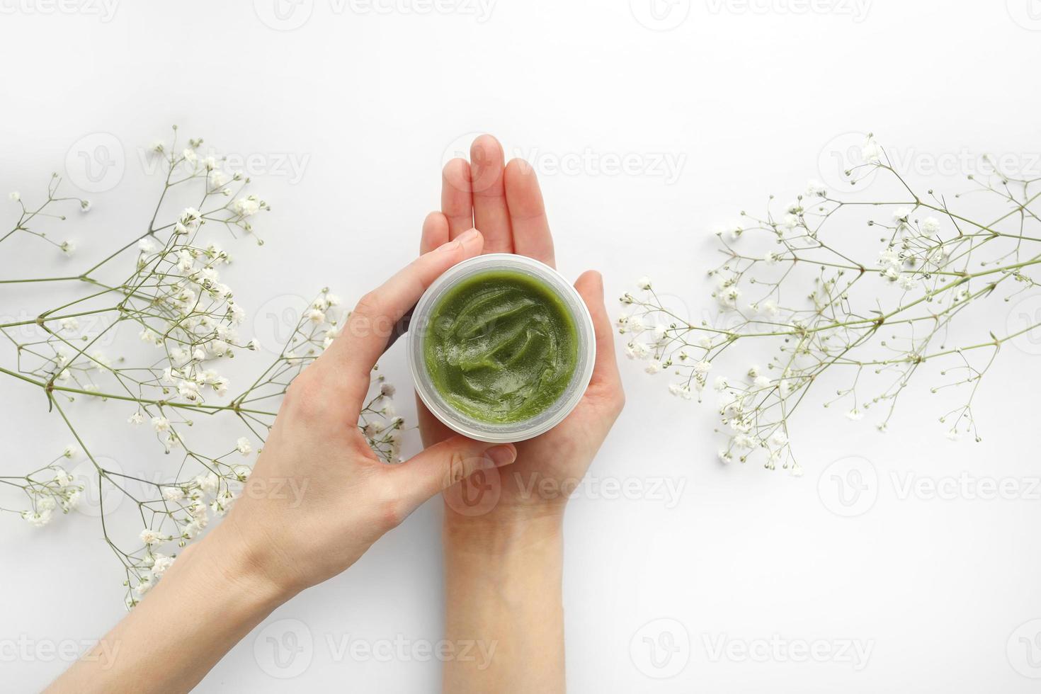 unga kvinnliga händer som håller en burk med grön naturlig kräm för ansikte eller kropp. ekologiska naturliga hudvårdsprodukter och blommor på vit bakgrund. förpackning av lotion eller grädde. skönhet kosmetisk hudvård koncept foto