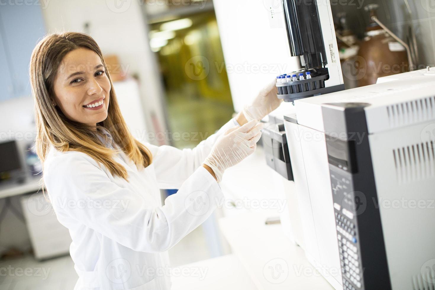 kvinnlig vetenskapsman i en vit labbrock sätter flaskan med ett prov för en analys på en gaskromatograf i biomedicinskt laboratorium foto