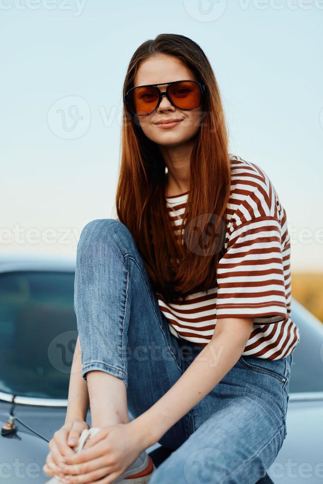en mode kvinna i eleganta solglasögon, en randig t-shirt och jeans sitter på de trunk av en bil och utseende på de skön natur av höst. resa livsstil foto