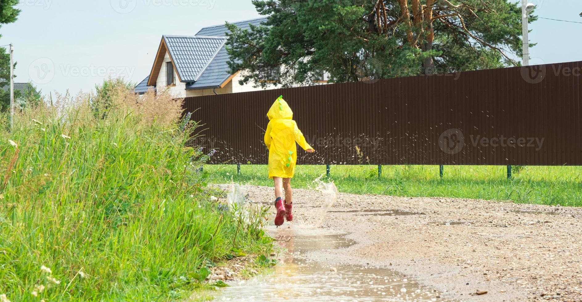 en flicka i röd sudd stövlar och en gul regnkappa kör genom vattenpölar efter en regn i de by. sommar tid, frihet, barndom foto