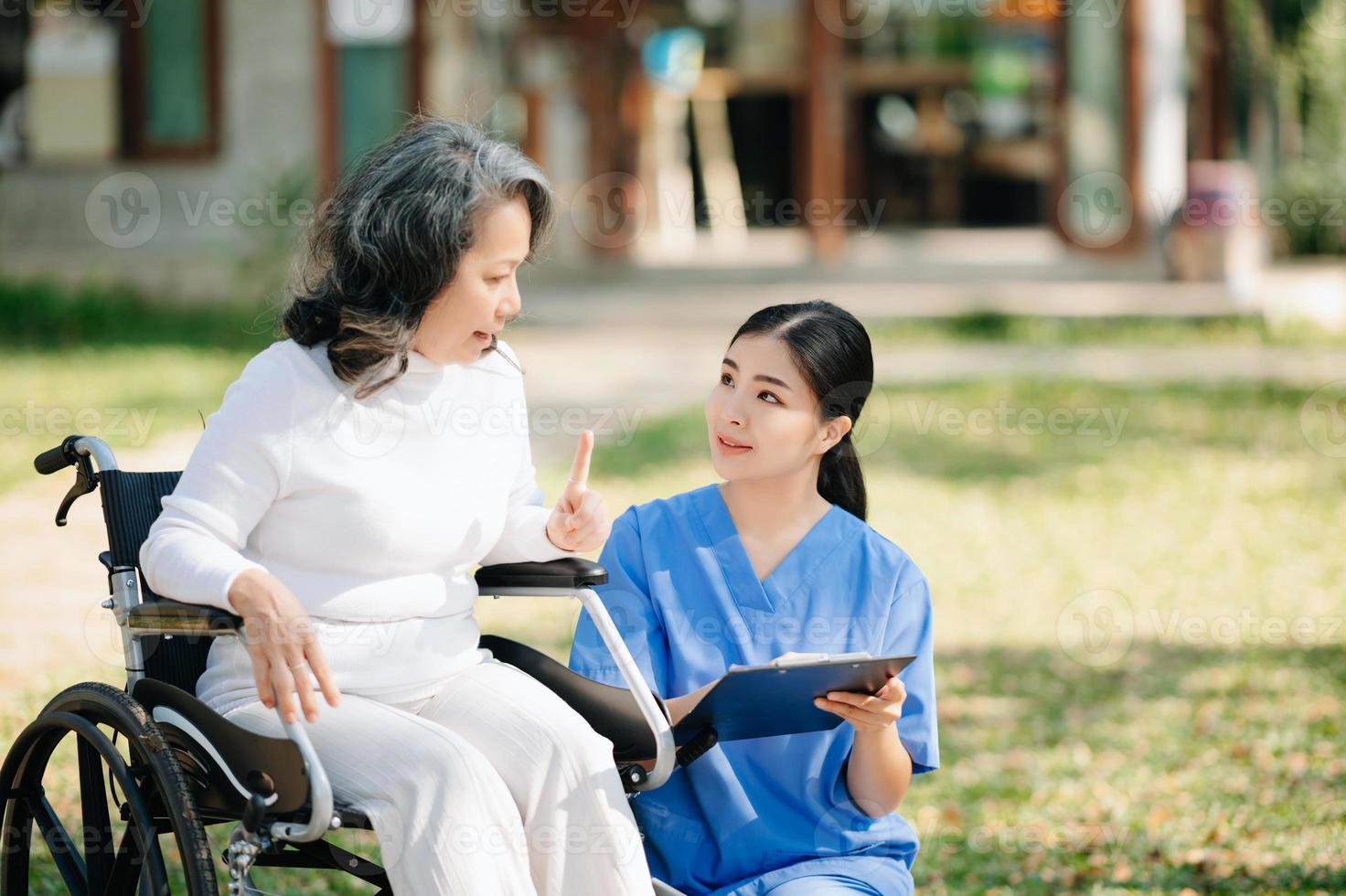 asiatisk försiktig vårdgivare eller sjuksköterska och de Lycklig patient i en rullstol är gående i de trädgård. till hjälp och uppmuntra och resten din sinne med grön natur. använda sig av en stetoskop till lyssna till din foto