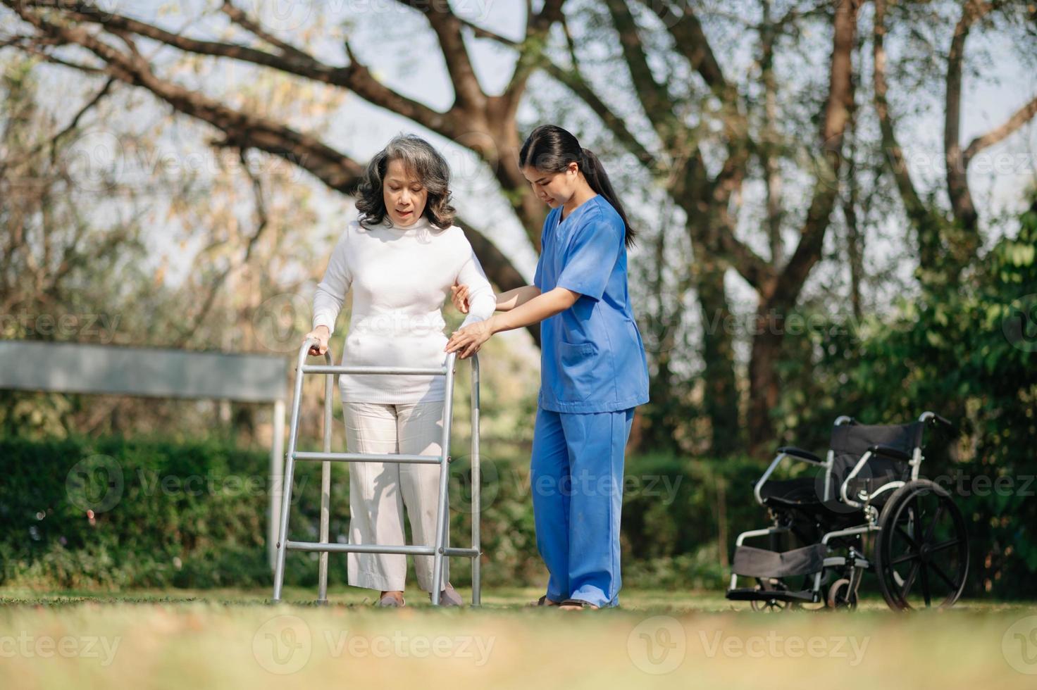 asiatisk försiktig vårdgivare eller sjuksköterska och de Lycklig patient i en rullstol är gående i de trädgård till hjälp och uppmuntra och resten din sinne med grön natur. hjälp Stöd själv till lära sig till promenad. foto