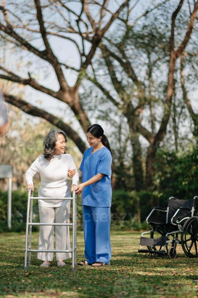 asiatisk försiktig vårdgivare eller sjuksköterska och de Lycklig patient i en rullstol är gående i de trädgård till hjälp och uppmuntra och resten din sinne med grön natur. hjälp Stöd själv till lära sig till promenad. foto