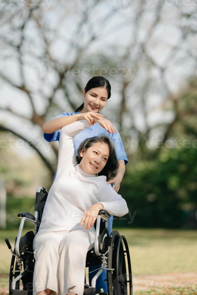 asiatisk försiktig vårdgivare eller sjuksköterska och de Lycklig patient i en rullstol är gående i de trädgård till hjälp och uppmuntra och resten din sinne med grön natur. hjälp Stöd själv till lära sig till promenad. foto