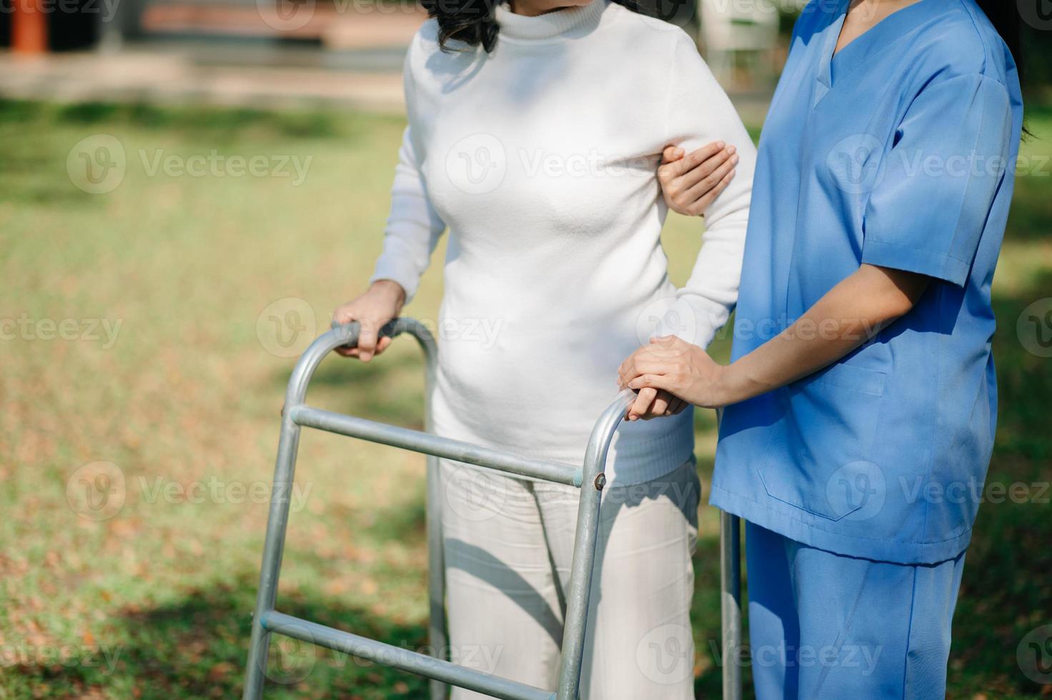 asiatisk försiktig vårdgivare eller sjuksköterska och de Lycklig patient i en rullstol är gående i de trädgård till hjälp och uppmuntra och resten din sinne med grön natur. hjälp Stöd själv till lära sig till promenad. foto