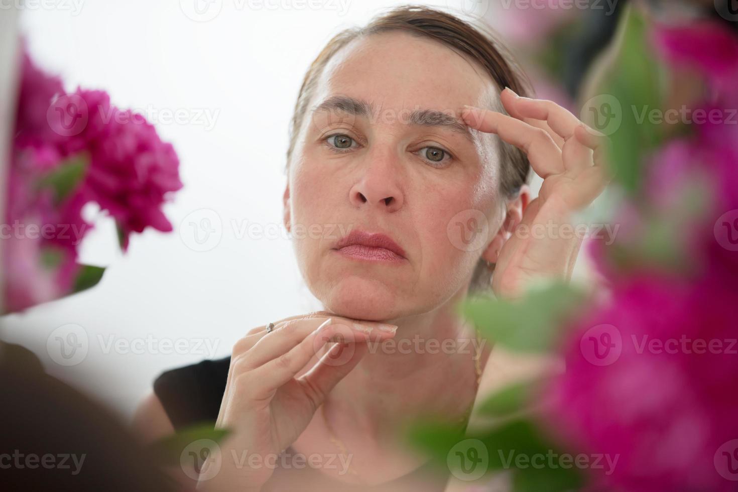 en mitten åldrig kvinna undersöker rynkor mot en bakgrund av blommor. vård för åldring hud. foto
