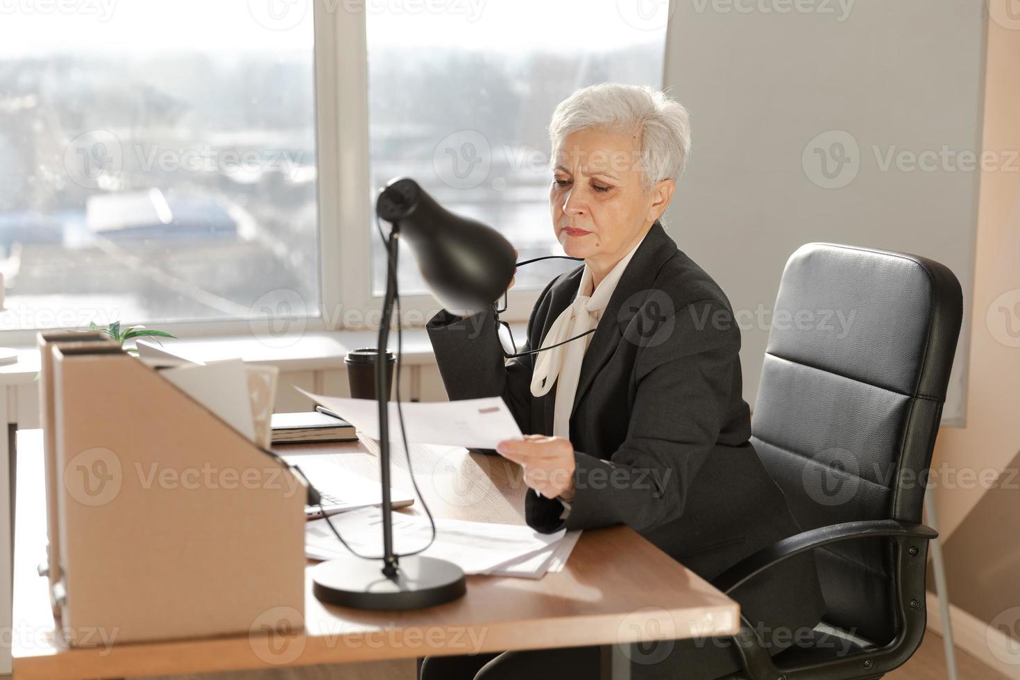 självsäker eleganta europeisk mitten åldrig senior kvinna läsning finansiell Rapportera dokumentation statistik. eleganta äldre mogna 60s grå håriga lady affärskvinna i kontor. chef ledare lärare. foto