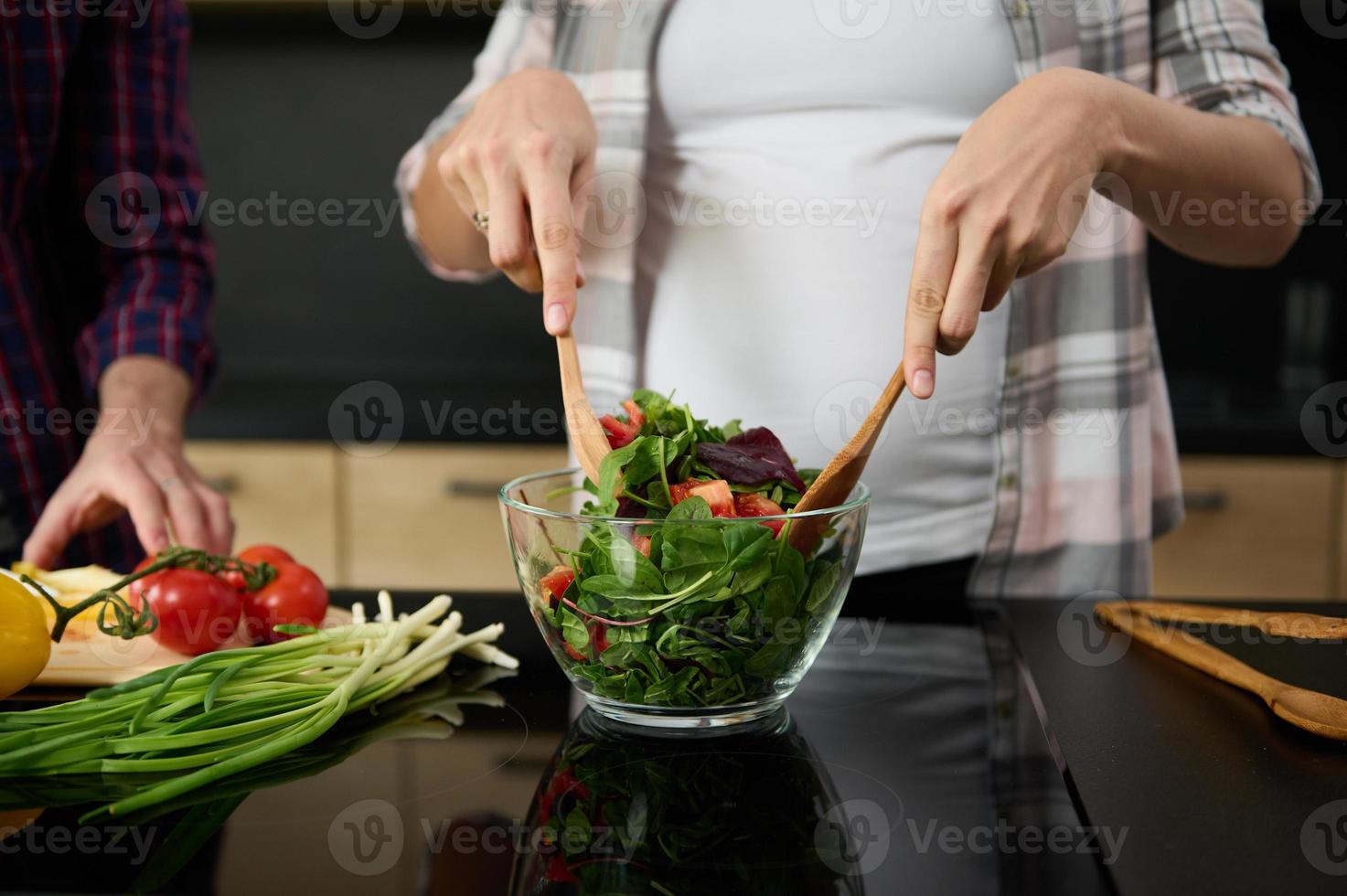 fokus på gravid kvinnas händer innehav trä- skedar och blandning Ingredienser i en glas skål, framställning utsökt friska sallad för middag i de kök ö Nästa till henne Make hackning grönsaker foto