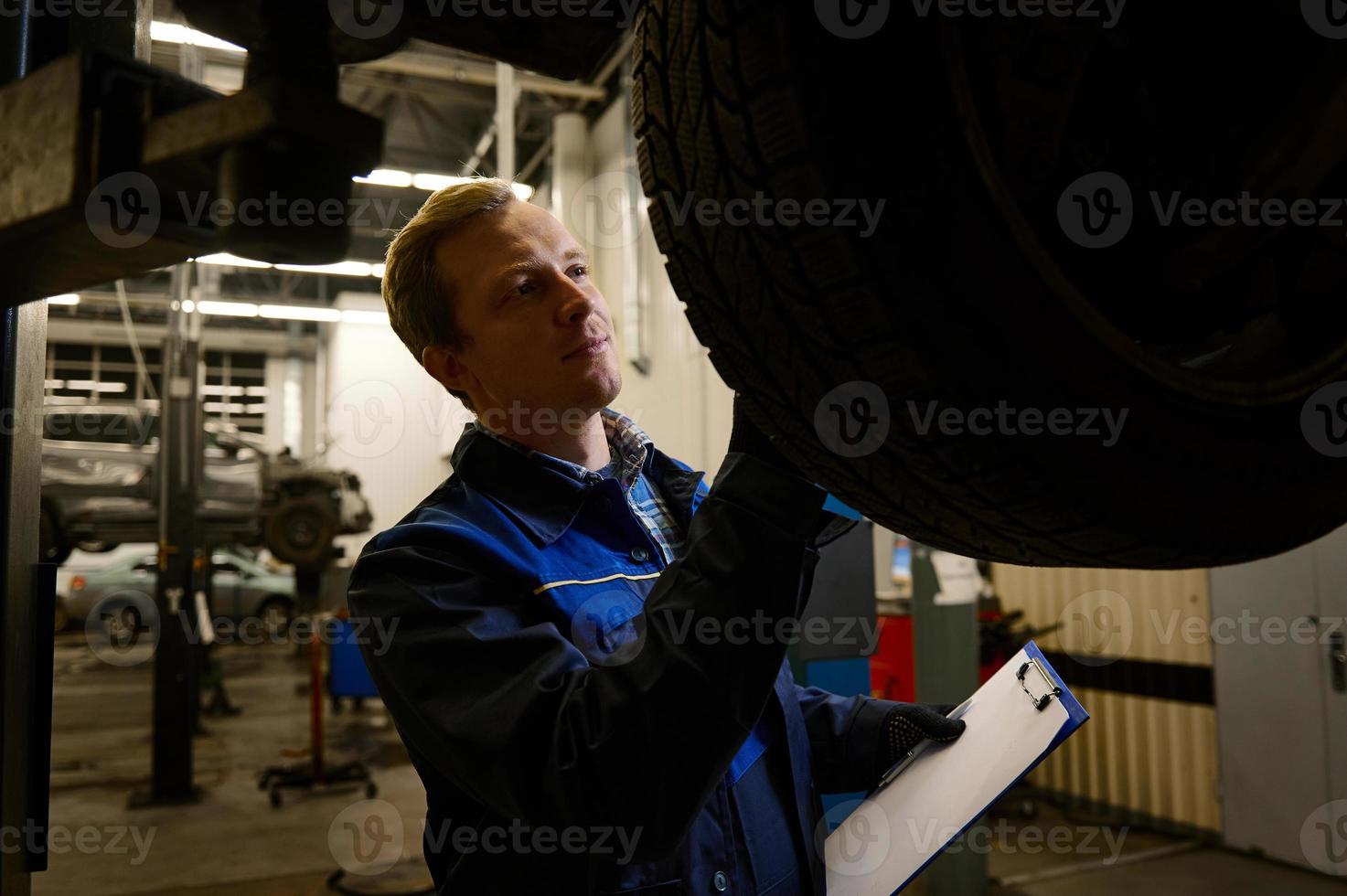 en ung behörig bil mekaniker inspekterar de bil Uppfostrad på de hiss för skada. begrepp av teknisk underhåll och bil service foto