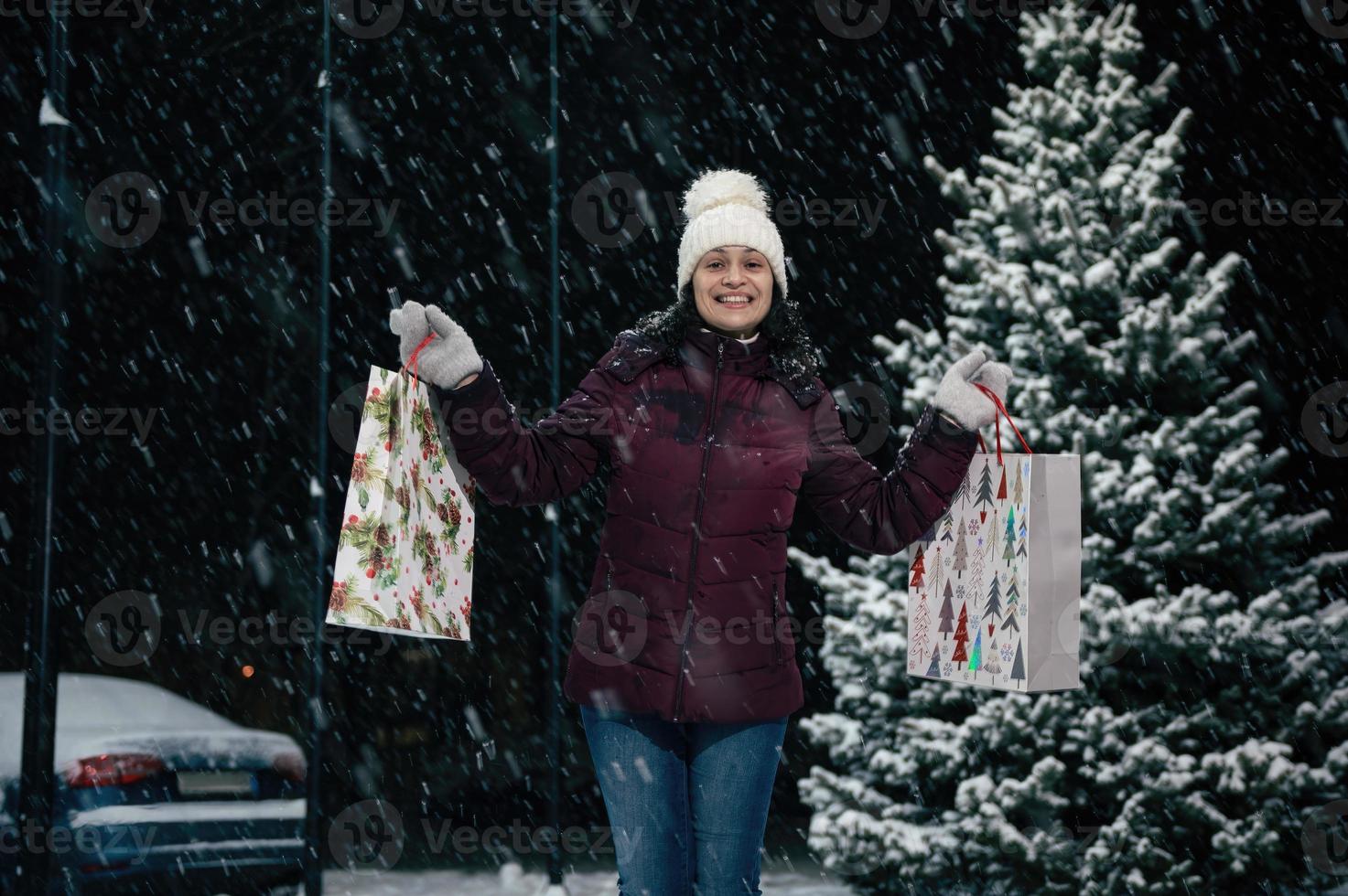 skön afrikansk amerikan kvinna i värma kläder, med handla påsar leende till de kamera på en snöig vinter- kväll foto