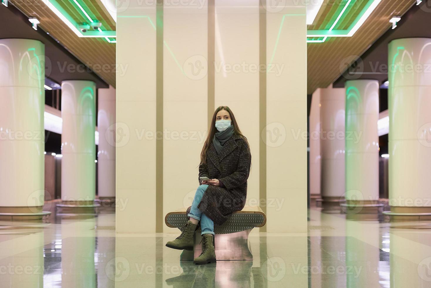 en kvinna i en medicinsk ansiktsmask väntar på ett tåg och håller en smartphone foto