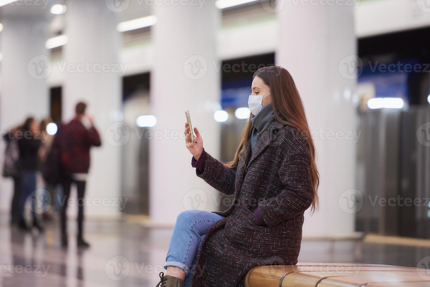 en kvinna i en medicinsk ansiktsmask väntar på ett tåg och håller en smartphone foto