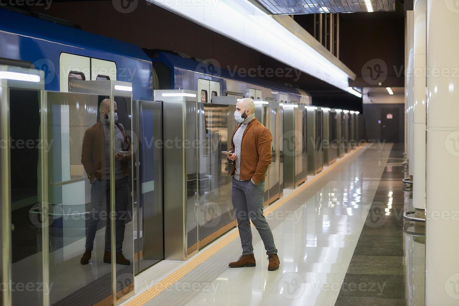 en man i ansiktsmask håller en smartphone medan han väntar på ett tunnelbanetåg foto