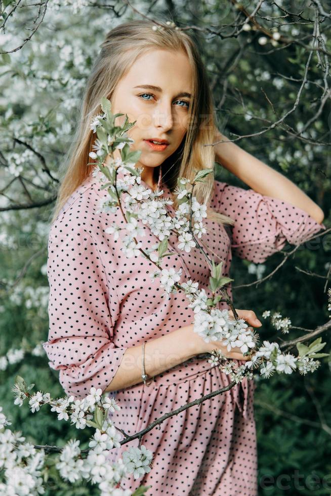 blond flicka på en vår promenad i de trädgård med körsbär blommar. kvinna porträtt, närbild. en flicka i en rosa polka punkt klänning. foto