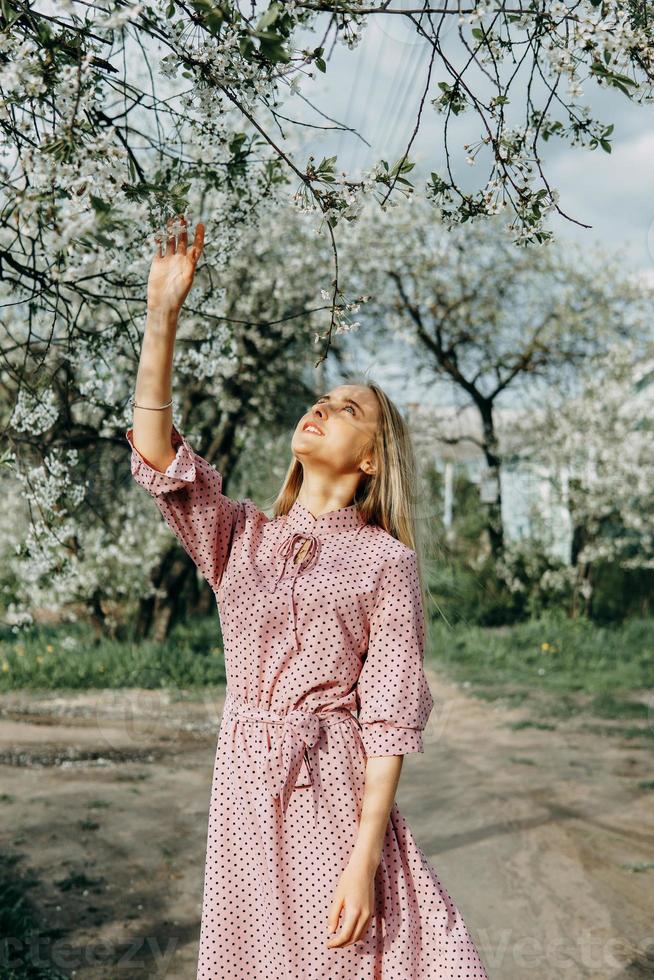 blond flicka på en vår promenad i de trädgård med körsbär blommar. kvinna porträtt, närbild. en flicka i en rosa polka punkt klänning. foto