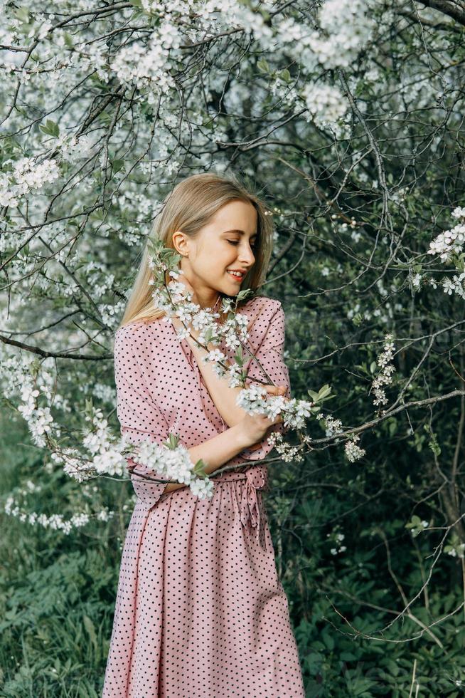 blond flicka på en vår promenad i de trädgård med körsbär blommar. kvinna porträtt, närbild. en flicka i en rosa polka punkt klänning. foto