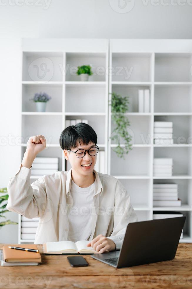 manlig studerande tar anteckningar från en bok på bibliotek, ung asiatisk Sammanträde på skrivbord håller på med uppgifter i högskola bibliotek foto