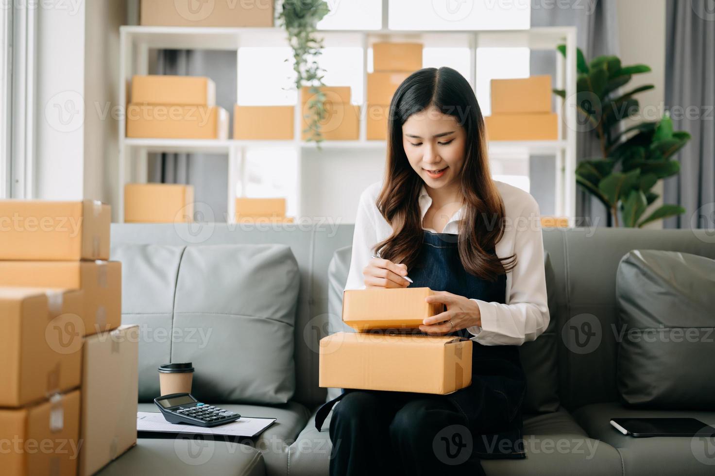 ung asiatisk företag ägare kvinna förbereda paket låda och stående kolla upp uppkopplad order för leverera till kund på läsplatta, bärbar dator handla uppkopplad begrepp. foto