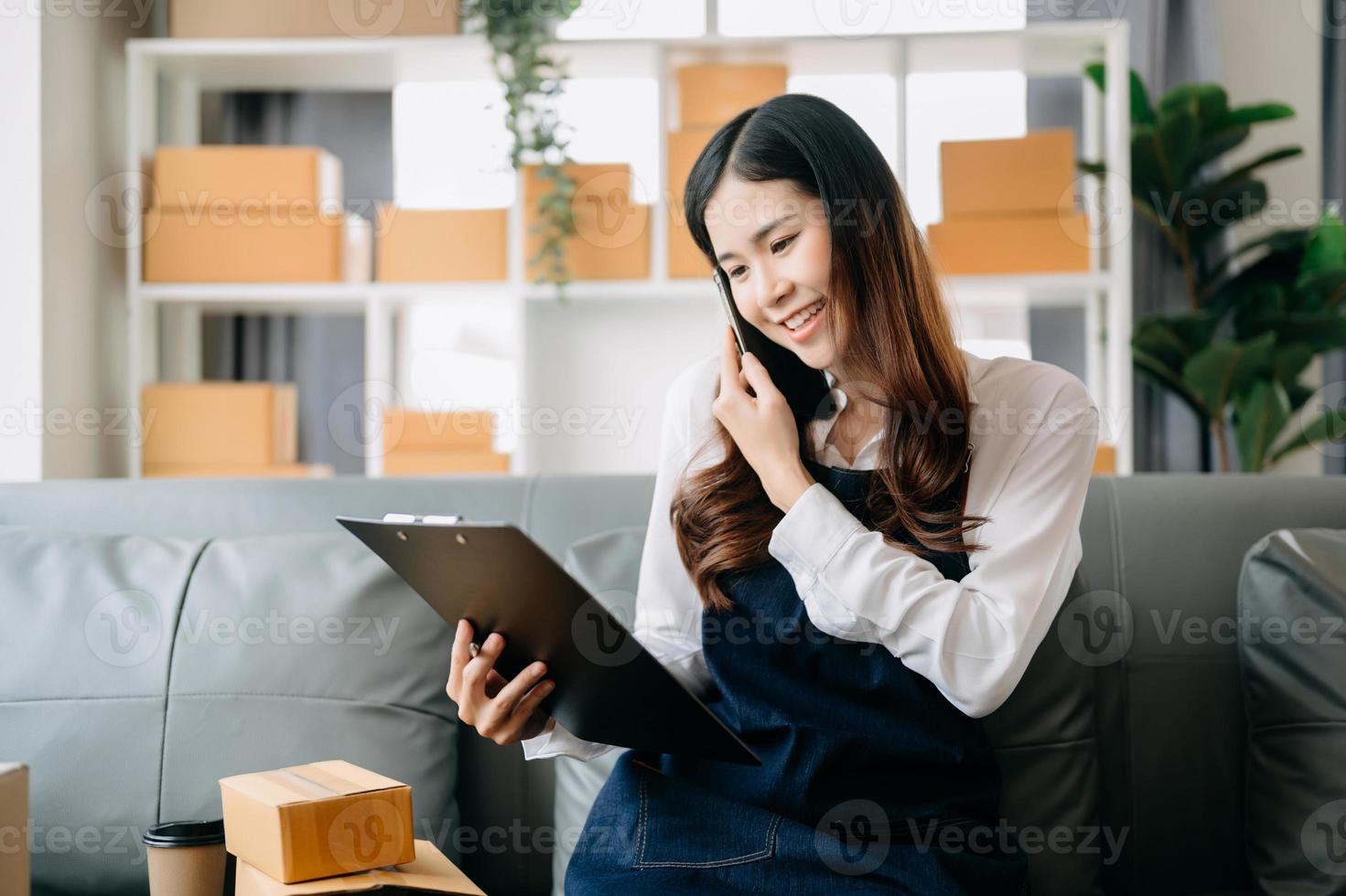 ung asiatisk företag ägare kvinna förbereda paket låda och stående kolla upp uppkopplad order för leverera till kund på läsplatta, bärbar dator handla uppkopplad begrepp. foto