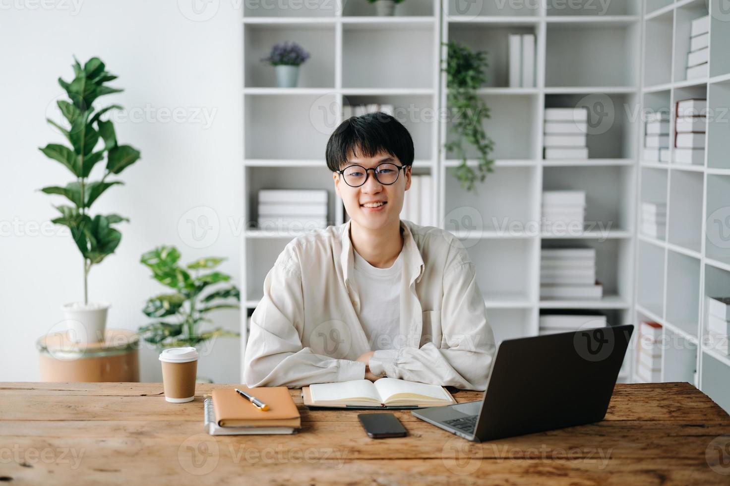 manlig studerande tar anteckningar från en bok på bibliotek, ung asiatisk Sammanträde på skrivbord håller på med uppgifter i högskola bibliotek foto