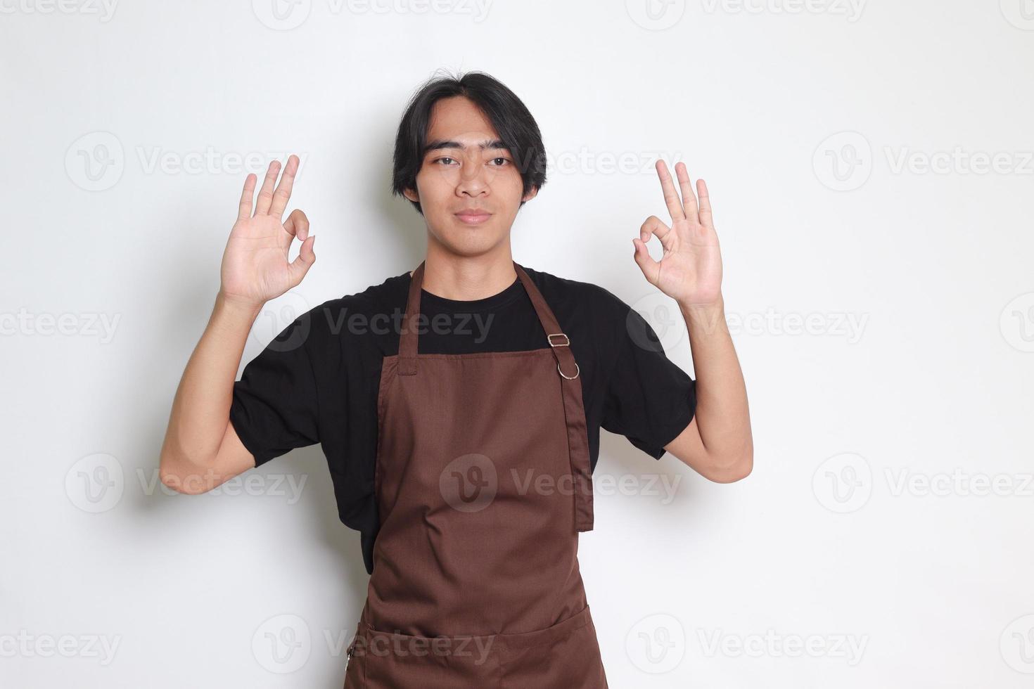 porträtt av attraktiv asiatisk Barista man i brun förkläde som visar ok hand gest och leende ser på kamera. reklam begrepp. isolerat bild på vit bakgrund foto