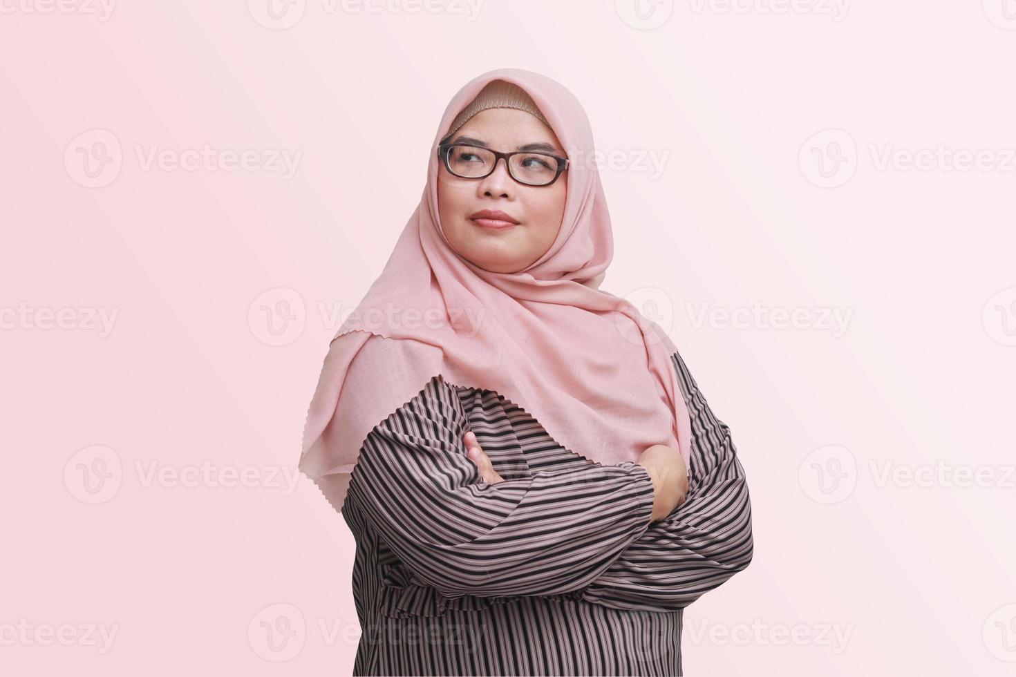 porträtt av självsäker asiatisk affärskvinna med hijab, förvaring vapen korsade och ser bort. isolerat bild på rosa bakgrund foto