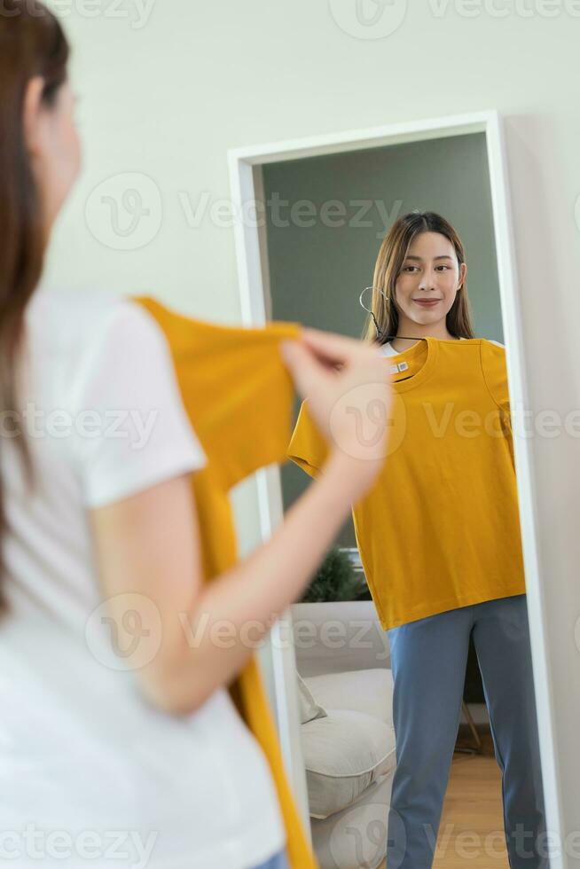 val av kläder, ingenting till ha på sig. attraktiv asiatisk ung kvinna, flicka ser in i spegel, Prova på verka, välja klänning, utrusta på galge i garderob på Hem. beslutar blus Vad till sätta på som ett foto